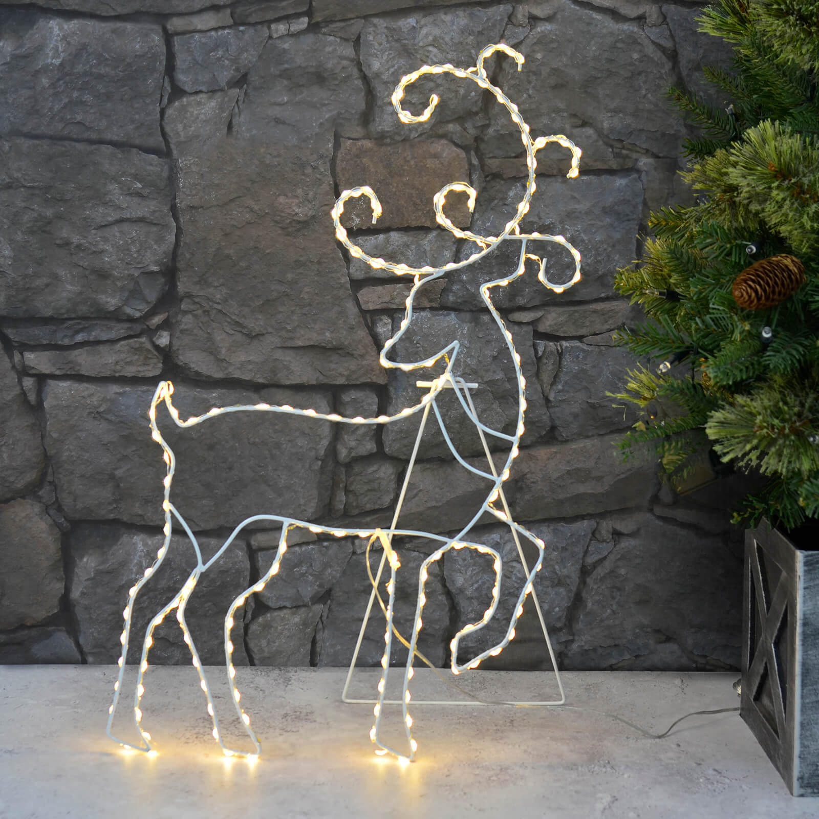 Christmas reindeer silhouette decoration lit by warm white LED lights on white wire frame on a path with stone wall and Christmas tree
